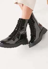 Botas de tobillo de charol negro brillante con cordones y cremallera lateral, combinadas con pantalones recortados de color beige claro sobre un fondo blanco.