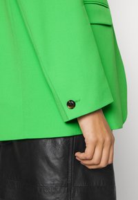 Blazer verde con una texture liscia, caratterizzato da un bottone nero sul polsino e dettagli delle tasche inclinati. Indossato su una gonna di pelle nera.