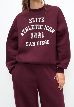 Kvinde iført vinrød sweatshirt og bukser med hvid tekst "Elite Athletic Icon 1991 San Diego" på sweatshirtens forside.