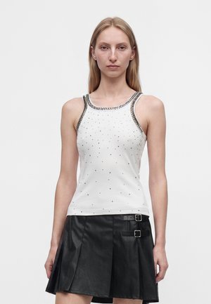 Femme portant un haut blanc sans manches avec des clous argentés et une bordure, assorti à une jupe plissée noire avec deux sangles à boucles.