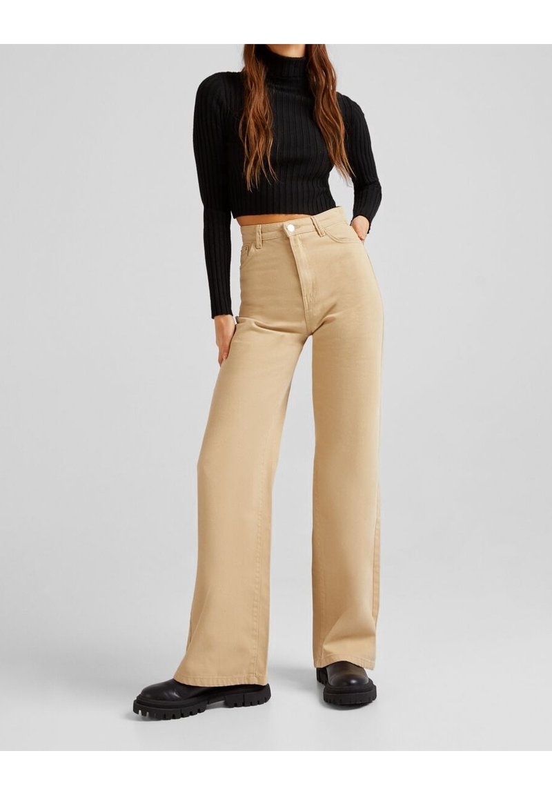 Pantalons larges beige en coton, avec une taille haute, une fermeture à bouton sur le devant, accompagnés d'un col roulé noir côtelé.