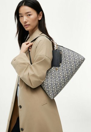 Giovane donna che indossa un trench beige e porta una grande borsa a mano con motivo, con una piccola pochette nera attaccata sulla spalla