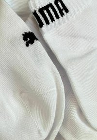 Weiße Socken aus Baumwolle mit einer gestrickten Textur. Merkmale schwarze Nähte mit dem Wort "ima" und einem kleinen geometrischen Akzent.