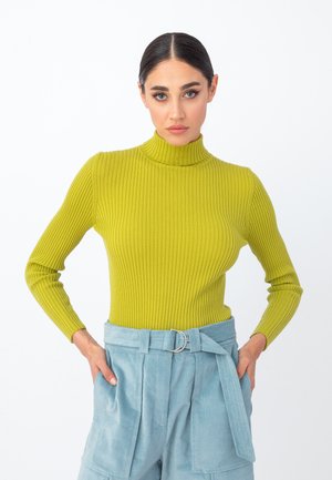 Rippstrick-Turtleneck-Pullover in leuchtendem Grün mit langen Ärmeln. Kombiniert mit hochgeschnittenen, hellblauen Hosen mit einem gebundenen Gürtel.