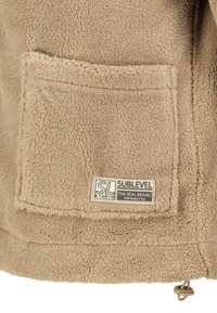 Beige Fleecejacke mit weicher Textur, ausgestattet mit einer Fronttasche und einem Logolabel. Verfügt über einen Kordelzug am Saum zur Anpassung.