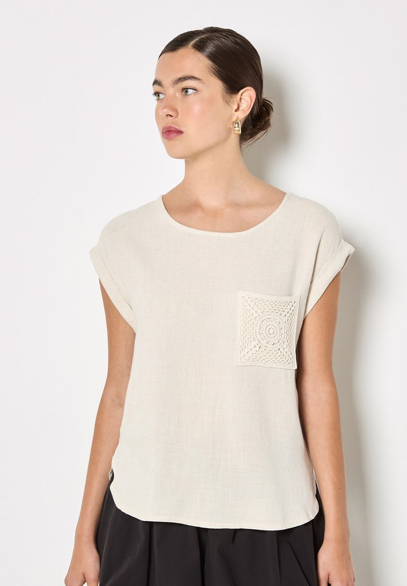 Jeune femme aux cheveux noirs attachés en chignon, portant un haut crème à manches courtes avec une poche avant en crochet et un pantalon noir, regardant vers la gauche.