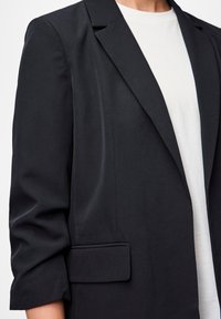 Blazer preto com um design estruturado, lapelas recortadas, mangas 3/4 e um único bolso frontal, combinado com uma camisa de cor clara.