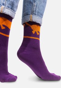 Fioletowe skarpetki o prążkowanej fakturze, z pomarańczowym wzorem dinozaurów i kontrastującą poziomą paskiem na górze. Widać mankiety dżinsów.