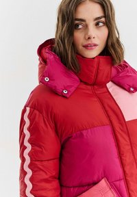Červená a růžová pufovaná bunda s vysokým límcem, patentkami, žebrovanými akcenty a designem s barevnými bloky s různými odstíny.