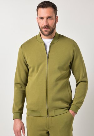 Man with beard wearing olive green zip-up jacket and matching pants, standing with one hand in pocket against plain background.