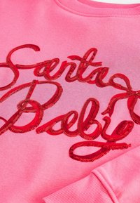 Tissu rose avec texte cursif rouge pailleté épelant "South" et "Bells" sur une manche de sweat partiellement visible.
