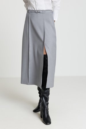 Jupe midi grise avec une fente avant, arborant une ceinture avec une boucle circulaire, texture lisse et design sur mesure ; associée à des bottes noires montantes jusqu'au genou.