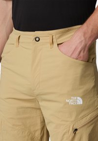 Tan shorts with a textured finish, featuring a button closure, front pockets, and a white logo on the left thigh.