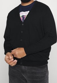 Svart kofta med V-ringad design, knäppning, långa ärmar och en slät textur; bärs över en mörk grafisk T-shirt.