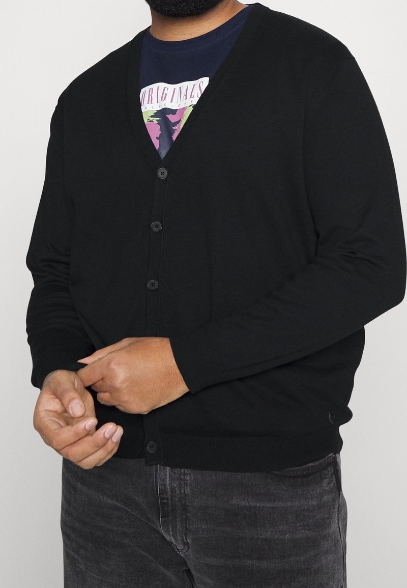 Svart kofta med V-ringad design, knäppning, långa ärmar och en slät textur; bärs över en mörk grafisk T-shirt.