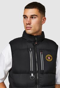 Zwarte gewatteerde vest met hoge kraag, ritssluiting aan de voorkant en twee borstzakken. Heeft een logo-opdruk en contrasterende witte ritsen.