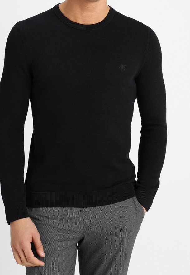 Homme portant un pull noir ajusté à manches longues et col rond ainsi qu’un pantalon gris texturé, debout avec une main dans la poche.