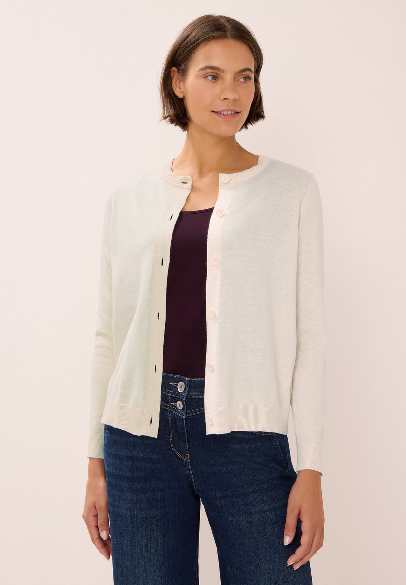 Vaalean kerma cardigan, joka on valmistettu pehmeästä materiaalista. Siinä on pyöreä kaula-aukko, napitus ja ribattu hihansuu. Päällä tumma paita ja farkut.