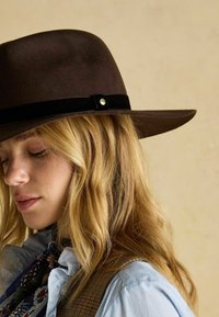 Pălărie maro din fetru cu bor larg, bandă din catifea neagră și detaliu cu nasture metalic. Textură moale și design structurat.