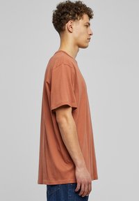 Hnědý bavlněný tričko s volnějším střihem, krátkými rukávy a kulatým výstřihem, vystavené na modelce v profilu proti šedému pozadí.