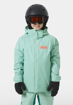 Kind in mintgrüner Skijacke und -hose, schwarzem Helm und Handschuhen, lächelnd mit orangefarbenen Skibrillen vor weißem Hintergrund.