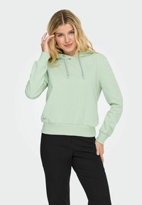 Felpa verde menta con tasca frontale, cordini, orlo a costine e un piccolo logo a forma di cuore. Abbinata a pantaloni neri per un contratto.