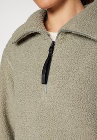 Pullover in pile verde chiaro con colletto a zip. Presenta una superficie texturizzata, una zip nera e cuciture grigie a contrasto.