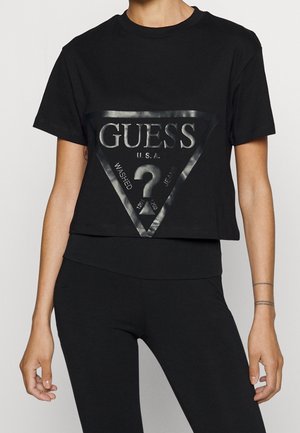 Osoba nosząca czarny krótki t-shirt z błyszczącym czarnym logo Guess USA i napisem wewnątrz trójkąta z przodu.