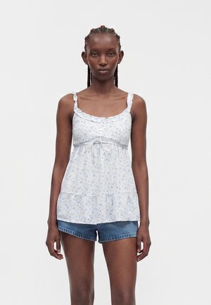 Vrouw met gevlochten haar die een witte mouwloze top met bloemenprint en ruches draagt en blauwe spijkershorts aan heeft, staand tegen een effen achtergrond.