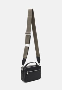 Sac bandoulière en synthétique noir avec poignée supérieure, présentant une sangle ajustable texturée crème et noire ainsi qu'une poche zippée à l'avant.