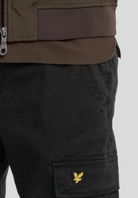 Brown zippered jacket features a ribbed waistband, paired with black cargo pants displaying a small yellow eagle logo on the pocket.