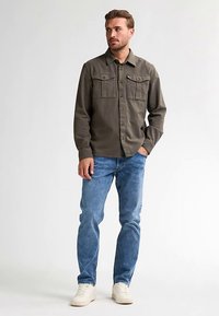 Olivengrøn skjorte med knapper og to brystlommer, kombineret med lyseblå jeans og hvide sneakers, med en afslappet pasform og blød tekstur.