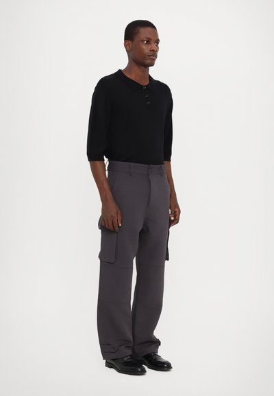 Zwarte poloshirt met drie knopen, mouwlengte tot halverwege de armen. Grijze cargobroek met zijzakken en een relaxte pasvorm. Zwarte schoenen.