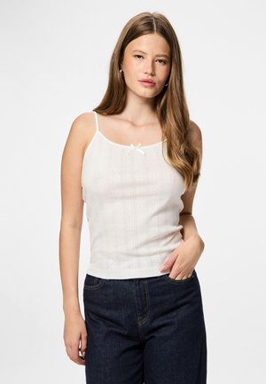 Camisole en tricot blanc avec des bretelles fines, ornée d'un petit nœud au niveau du cou et d'un motif rayé texturé. Accompagnée de jeans bleu marine.