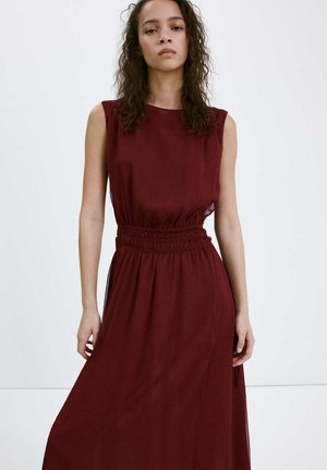 Mujer con cabello rizado y castaño que lleva un vestido midi sin mangas de color rojo oscuro con cintura fruncida, de pie frente a un fondo blanco liso.