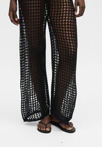 Jambes portant un pantalon ample noir en maille ajourée laissant voir la peau en dessous et des sandales tongs noires sur pieds nus.