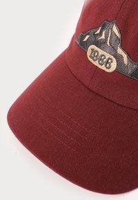 Burgundi sapka strukturált anyaggal, hímzett hegydesignnal és "1966" jelvénnyel bézs és fekete színben az elején.