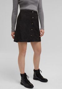 Zwarte suede rok met een uitlopende vorm, voorzien van zes knopen aan de voorkant. Gepaard met zwarte veterschoenen en een getailleerd grijs longsleeve shirt.