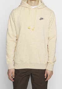 Felpa beige in tessuto texture, con tasca frontale, cappuccio con cordino e punte gialle, e un piccolo logo Nike nero sul petto.