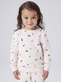 Jeune fille aux longs cheveux bruns portant un sweat-shirt rose clair et un pantalon avec des imprimés de cerises bordeaux, souriant à la caméra sur un fond uni.