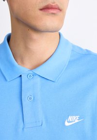 Svetlomodrá polo tričko vyrobené z textúrovanej tkaniny. Má klasický golier, dva gombíky a biele logo Nike na ľavej strane hrude.