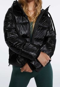 Svart vadderad jacka med glänsande, quiltad yta; har huva, dragkedja fram och elastiska muddar. Bärs över en mörkgrön outfit.