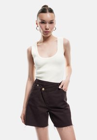 Top sin mangas blanco con relieve y bordes ondulados, combinado con pantalones cortos de mezclilla marrón oscuro con dobladillo deshilachado y bolsillos laterales.