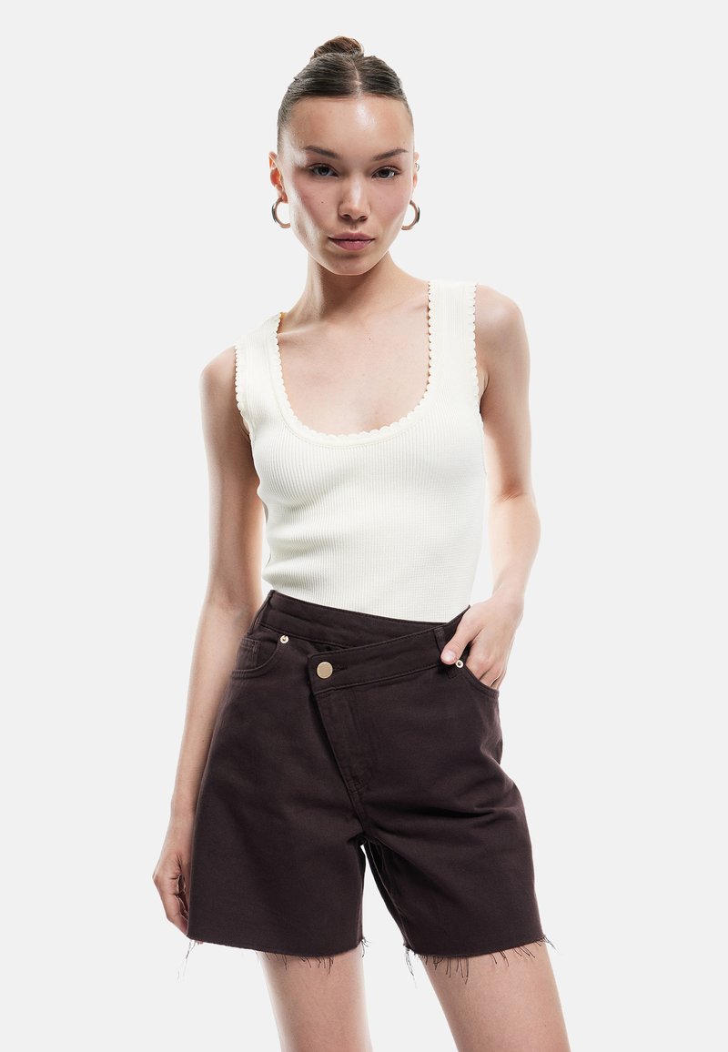 Top sin mangas blanco con relieve y bordes ondulados, combinado con pantalones cortos de mezclilla marrón oscuro con dobladillo deshilachado y bolsillos laterales.