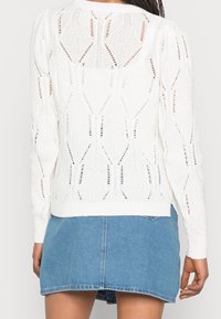 Biały dziergany sweter z wzorzystym designem i ażurowymi detalami. Ściągacze na mankietach i na dole, w połączeniu z dżinsową spódnicą.