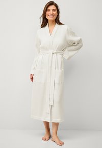 Robe longue en tricot gaufré de couleur crème avec une ceinture, des manches larges et deux poches avant, présentant un tissu texturé et une coupe à mi-genoux.