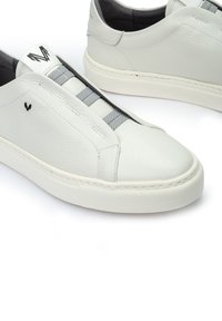 Zapatillas de cuero blanco con acabado texturizado, bandas elásticas gris para el cierre, diseño minimalista y suela de goma blanca. Detalle del logo en negro presente.