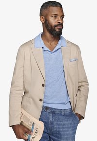 Mężczyzna z brodą, ubrany w beżową marynarkę, jasnoniebieskie polo i dżinsy, trzymający złożoną gazetę w lewej ręce, patrzący na prawo z neutralnym wyrazem twarzy.