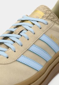 Μπεζ σουέντ αθλητικό παπούτσι με ανοιχτό μπλε ρίγες, διαθέτει υφασμάτινες λεπτομέρειες, χρυσό λογότυπο και κορδόνια στο ίδιο χρώμα. Εφοδιασμένο με καφέ καουτσούκ σόλα.