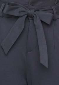 Marinblått tyg med en knytning i midjan och sidofickor. Slät textur med lätt stretch, med en ren design och inga synliga mönster.